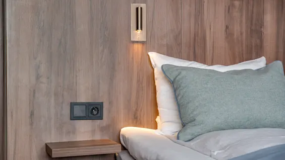 Bed with light gray pillow resting on white sheets. Wall-mounted reading light shines above a small shelf. Wooden panel backdrop gives a warm, cozy atmosphere. 