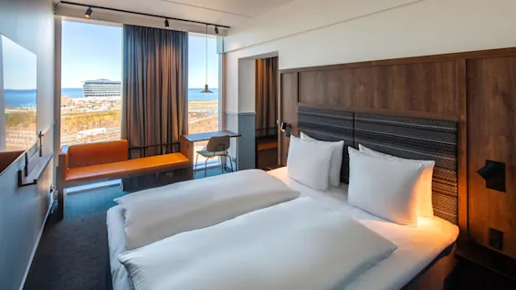 En pent, oppredd seng med hvitt sengetøy står i et moderne hotellrom med sengegavl av tre. Et vindu med utsikt over sjøen og bygninger slipper inn naturlig lys.