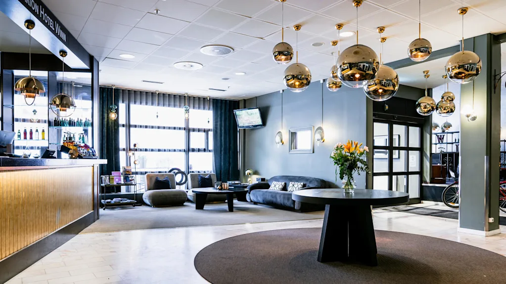 Ett hotellobby med modern inredning har ett runt bord med blommor, hängande sfäriska lampor och en mysig sittgrupp. Text: "Clarion Hotel Winn" på receptionsdisken.