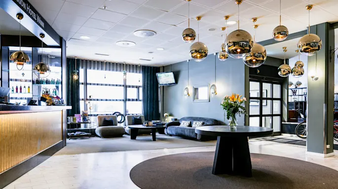 En hotellobby med moderne innredning har et rundt bord med blomster, hengende sfæriske lamper, og et koselig sitteområde. Tekst: "Clarion Hotel Winn" på resepsjonsdisken.