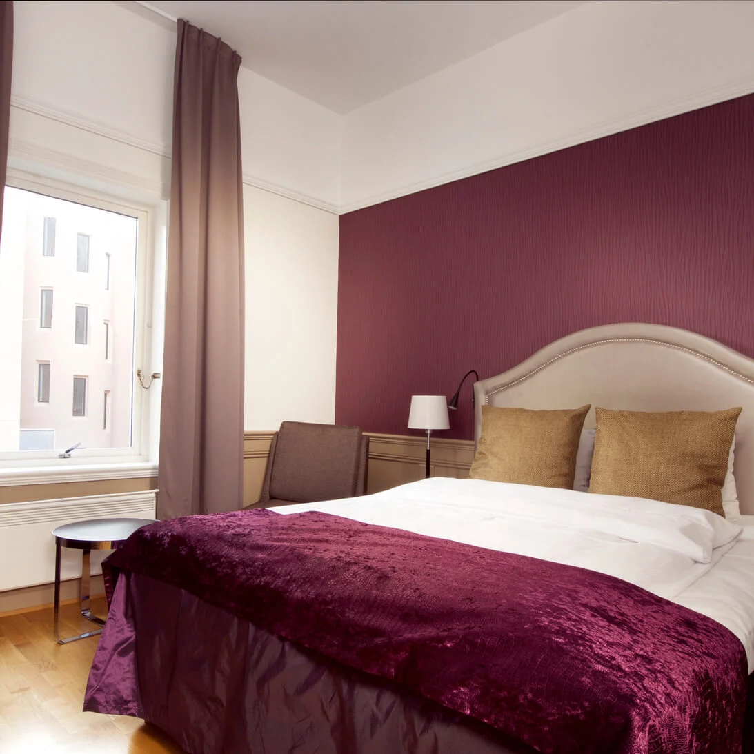 En pent oppredd seng med et lilla kast i et soverom med en maroon aksentvegg, vindu og møbler.
