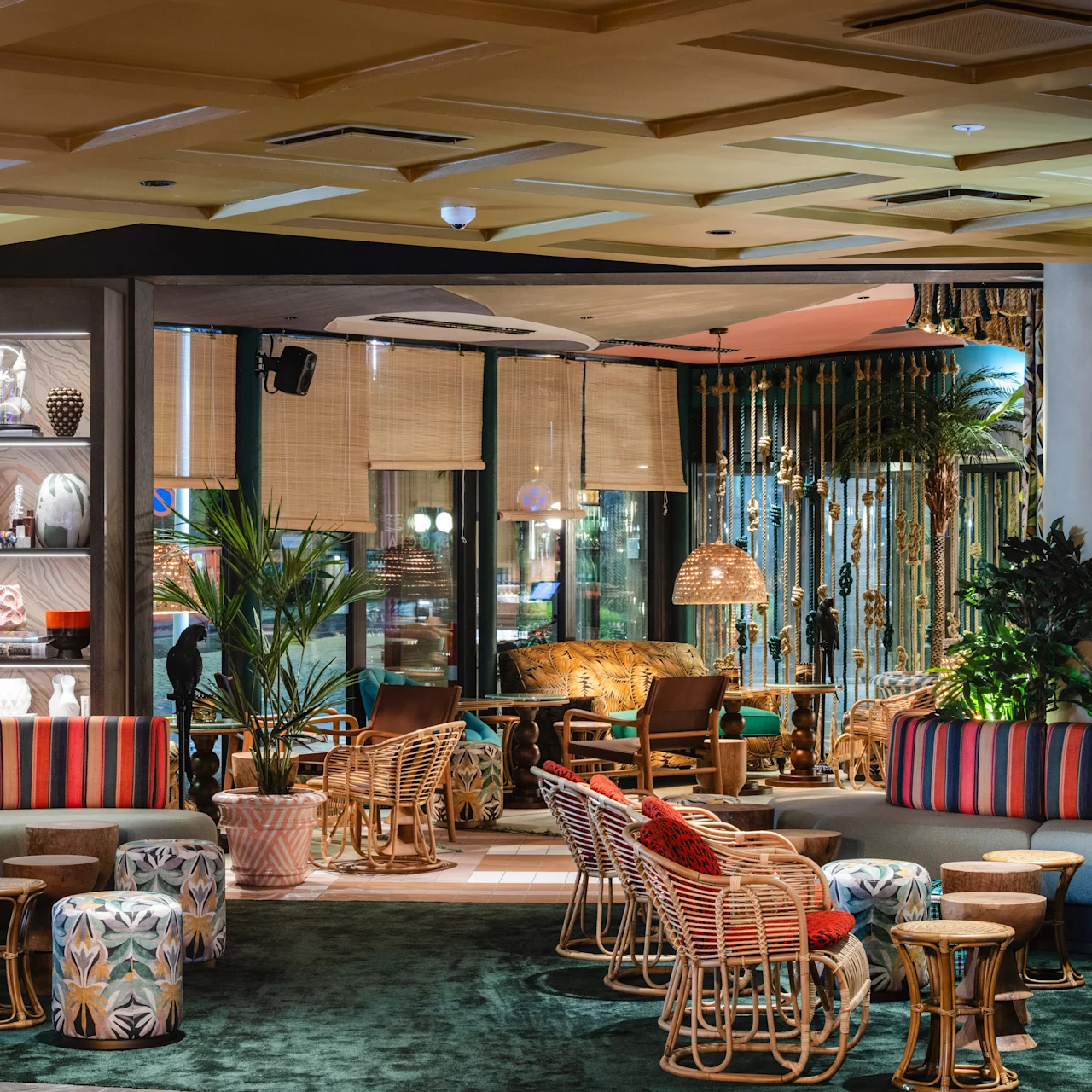 Colorful seating arrangement with striped cushions, surrounded by tropical plants and wicker furniture. Soft lighting from hanging lamps creates a cozy, stylish atmosphere inside a modern lounge setting.