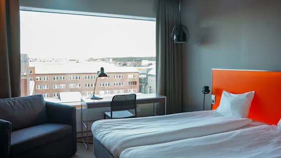 En seng med et orange sengegærde; et skrivebord ved siden af, med en lampe, vender mod et stort vindue med udsigt over en by med murstensbygninger.