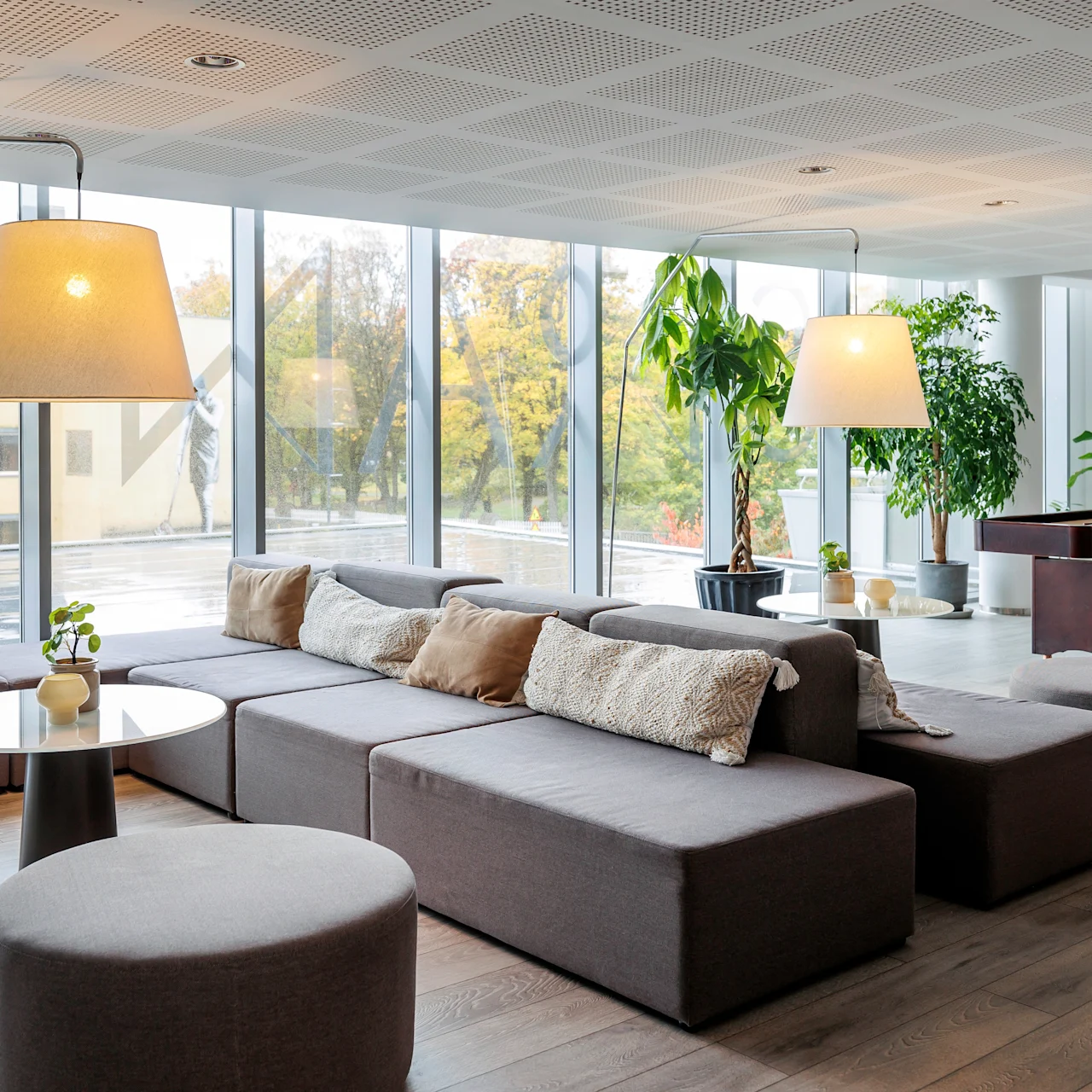 A modern lounge with gray modular sofas and large pendant lamps. Potted plants soften the space, and expansive windows reveal an outdoor view. A shuffleboard table stands nearby.