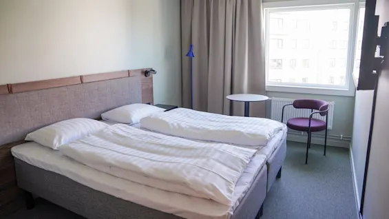 En dobbeltseng er pent oppredd mot en lys grå vegg, ved siden av et vindu. Et lite rundt bord og en lilla stol står like ved, med lukkede beige gardiner.