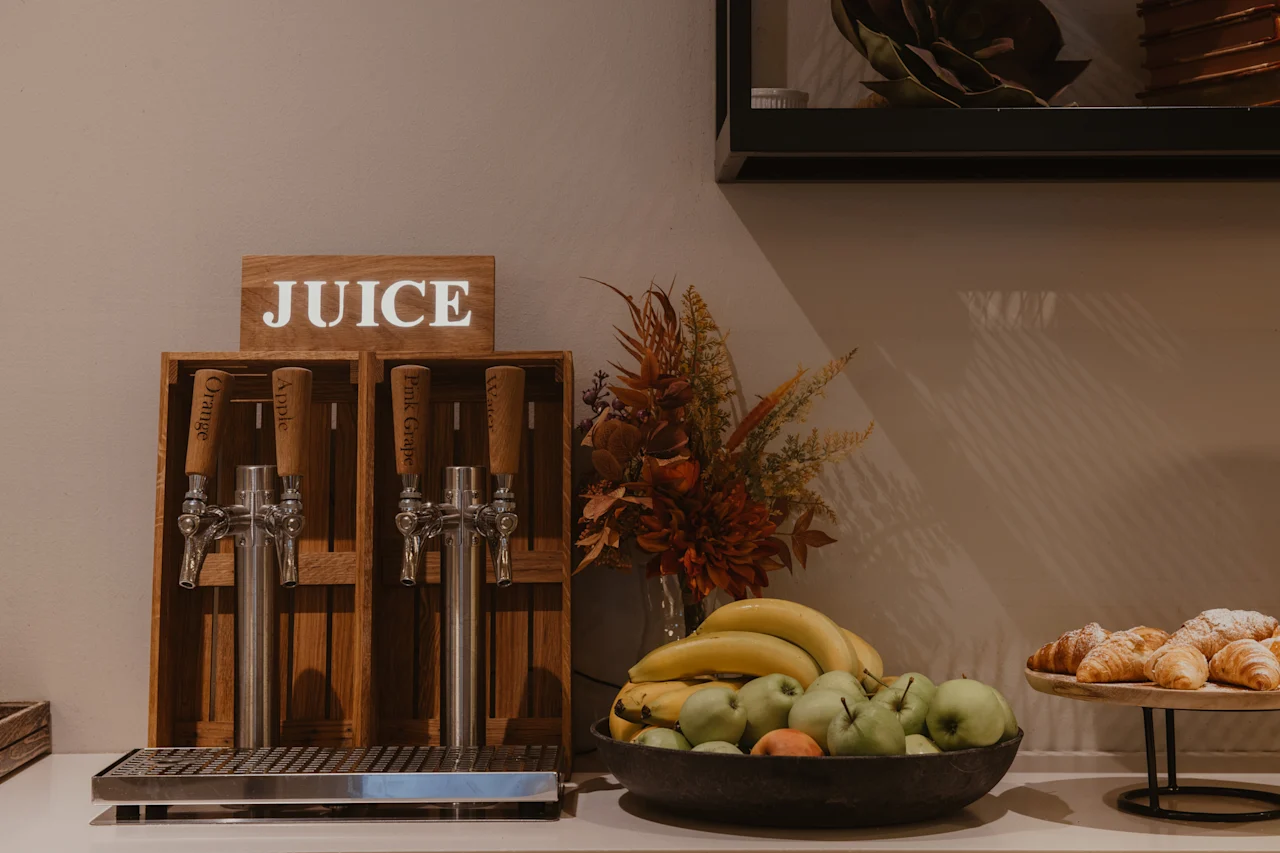 A self-serve juice station with options for orange, apple, pink grape, and water, alongside fresh fruit and croissants.