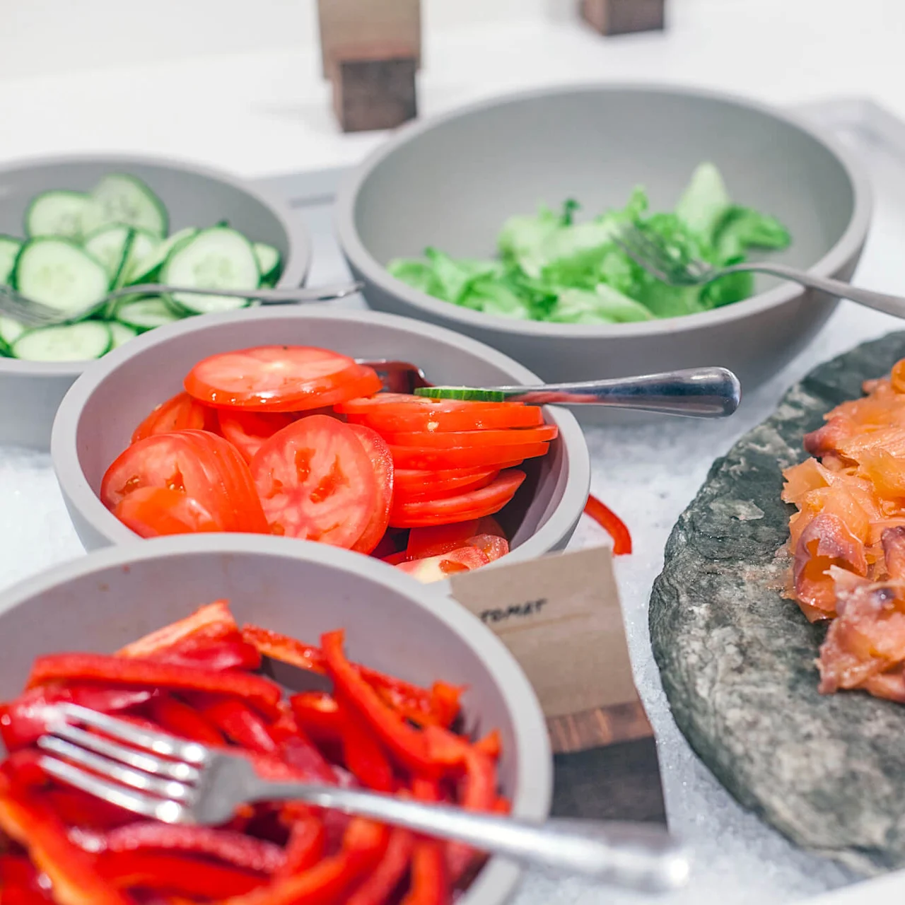 Skiver av laks ligger på en steinplate, akkompagnert av sitronbåter, med boller av skiver av tomater, agurker, salat og rød paprika i nærheten.