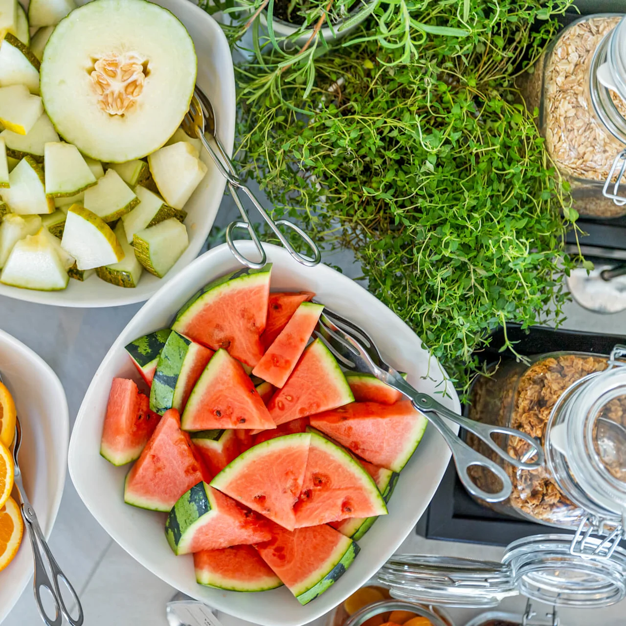 Fresh fruit slices in bowls, with watermelon and oranges, next to growing cress and jars of oats on a kitchen countertop.