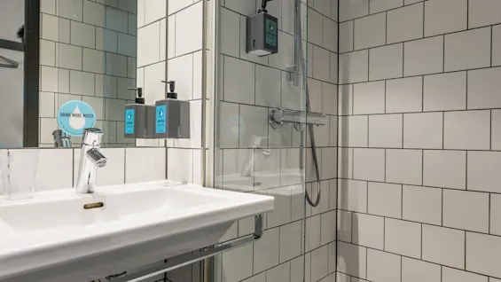 Tvättställ på en vit kaklad vägg med en spegel ovanför, flankerad av tvåltvålar märkta "LUXURY HAND WASH," bredvid en glasdusch med duschmunstycke.
