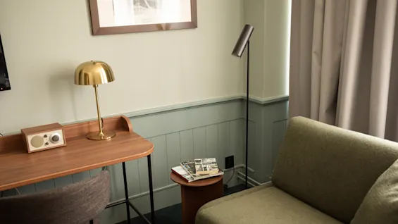 Ett mysigt hörn med ett skrivbord i trä, guldig lampa, retro radio, grå stol, grön soffa, sidobord med tidningar och mjuka gardiner i ett dovt belyst rum.