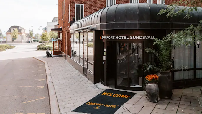 Inngang til Comfort Hotel Sundsvall med sort markise og potteplanter; en velkomstmatte har påskriften "WELCOME COMFORT HOTEL."