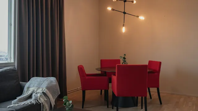 En matplats med ett runt bord omgivet av röda stolar. Hängande taklampor lyser upp utrymmet. I närheten kompletterar en grå soffa med en filt och ett litet bord det mysiga rummet.