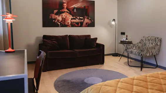 Brun soffa under ett stort fotografi, flankerad av en zebramönstrad stol och ett sidobord med vinglas, i ett mysigt rum med varm belysning och en röd skrivbordslampa.