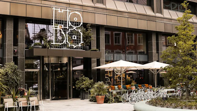Hotel facade displays geometric Hobo signage above entrance, with potted plants and outdoor seating under umbrellas on a city street.