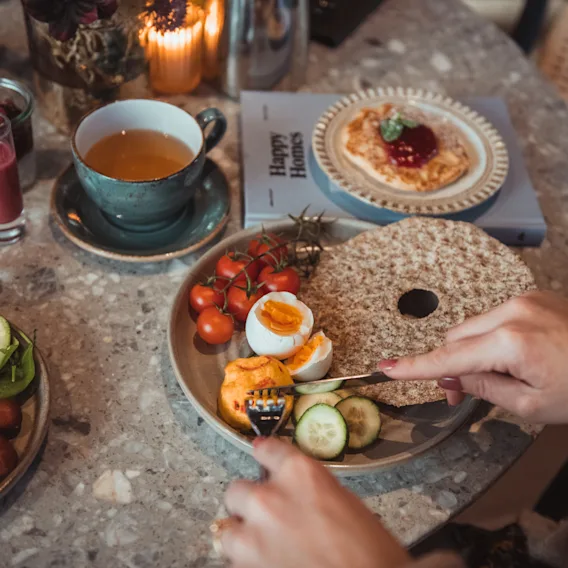 En person skjærer et kokt egg på en tallerken med tomater, agurker og knekkebrød på et trebord. I nærheten er det te, juice og en bok med tittelen "Happy Food."