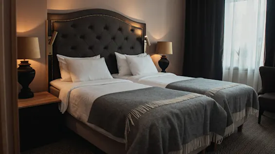 Two neatly made twin beds with gray throws and white pillows, flanked by dark nightstands with lamps, in a softly lit room featuring a large window with sheer curtains.