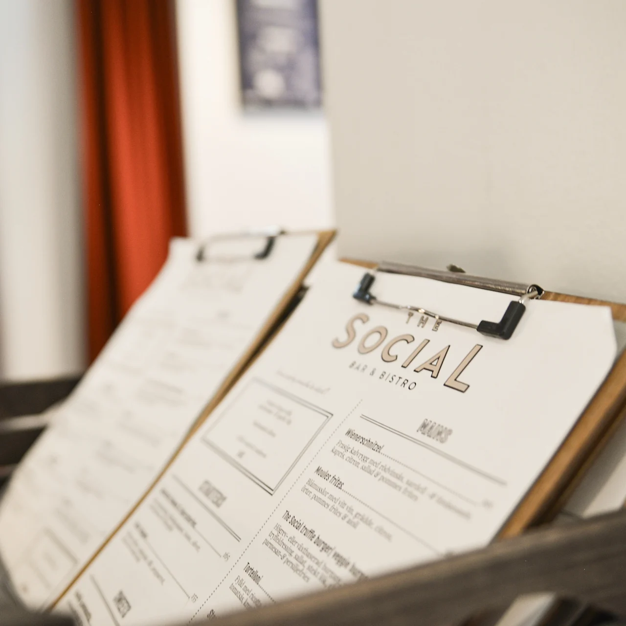 Menuklips hviler i en trækasse i en bistro. Tekst lyder "THE SOCIAL BAR & BISTRO" og "MENU." Sløret baggrund med rød gardin og hvid væg.