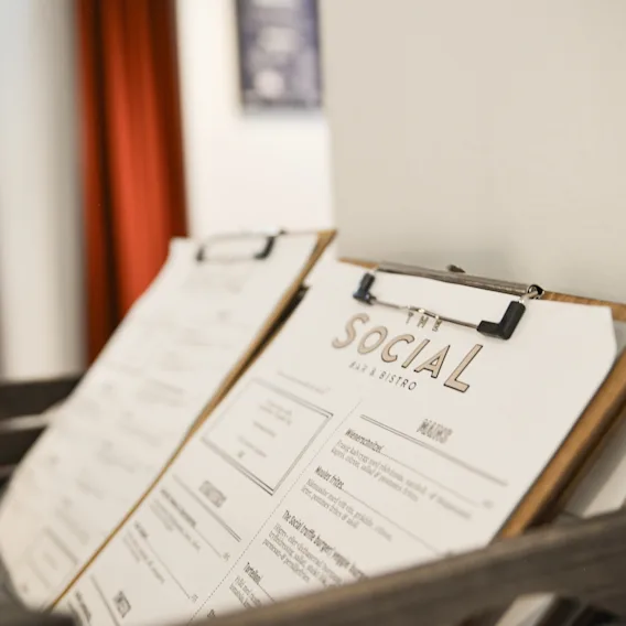 Clipboard menus rest in a wooden holder in a bistro. Text reads "THE SOCIAL BAR & BISTRO" and "MENU." Blurred background includes a red curtain and a white wall.