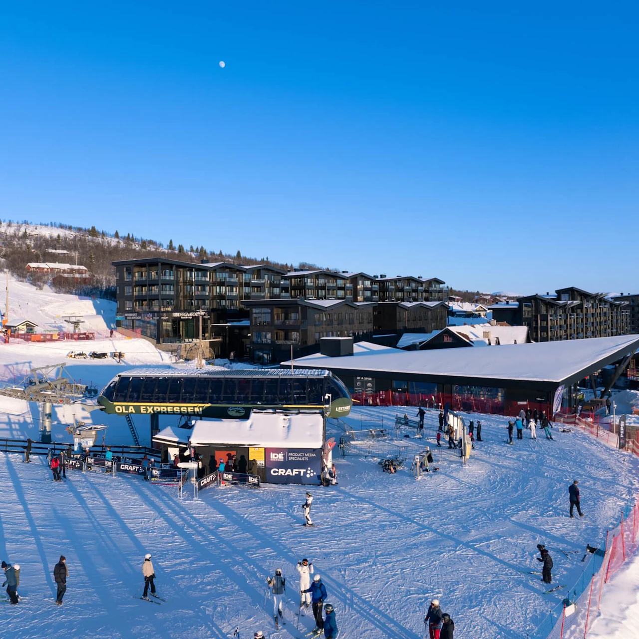 En skiliftstation mærket "OLA EKSPRESEN" med folk, der samles og står på ski i et snedækket område. Tæt på er moderne bygninger under en klar blå himmel.