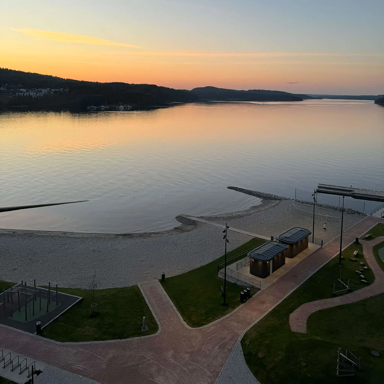 Sunset over a calm lake with a sandy beach, grassy park area, small buildings, pathways, and distant forested hills creating a tranquil landscape.