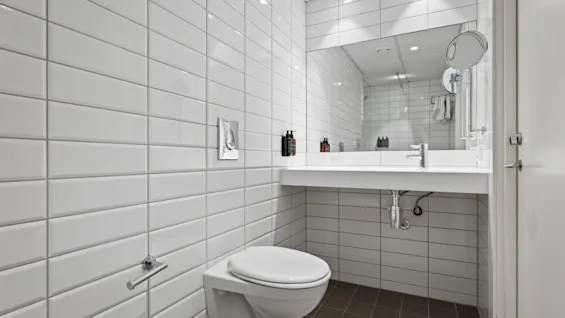 Ett rent och modernt hotellbadrum med vita kaklade väggar, en vägghängd toalett och ett handfat med en stor spegel.