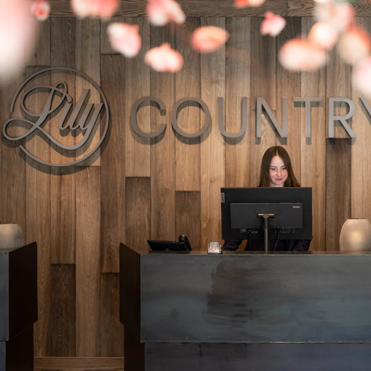 En kvinna jobbar vid en receptionsdisk med skyltningen "Lily Country Club" på träpanel i bakgrunden.