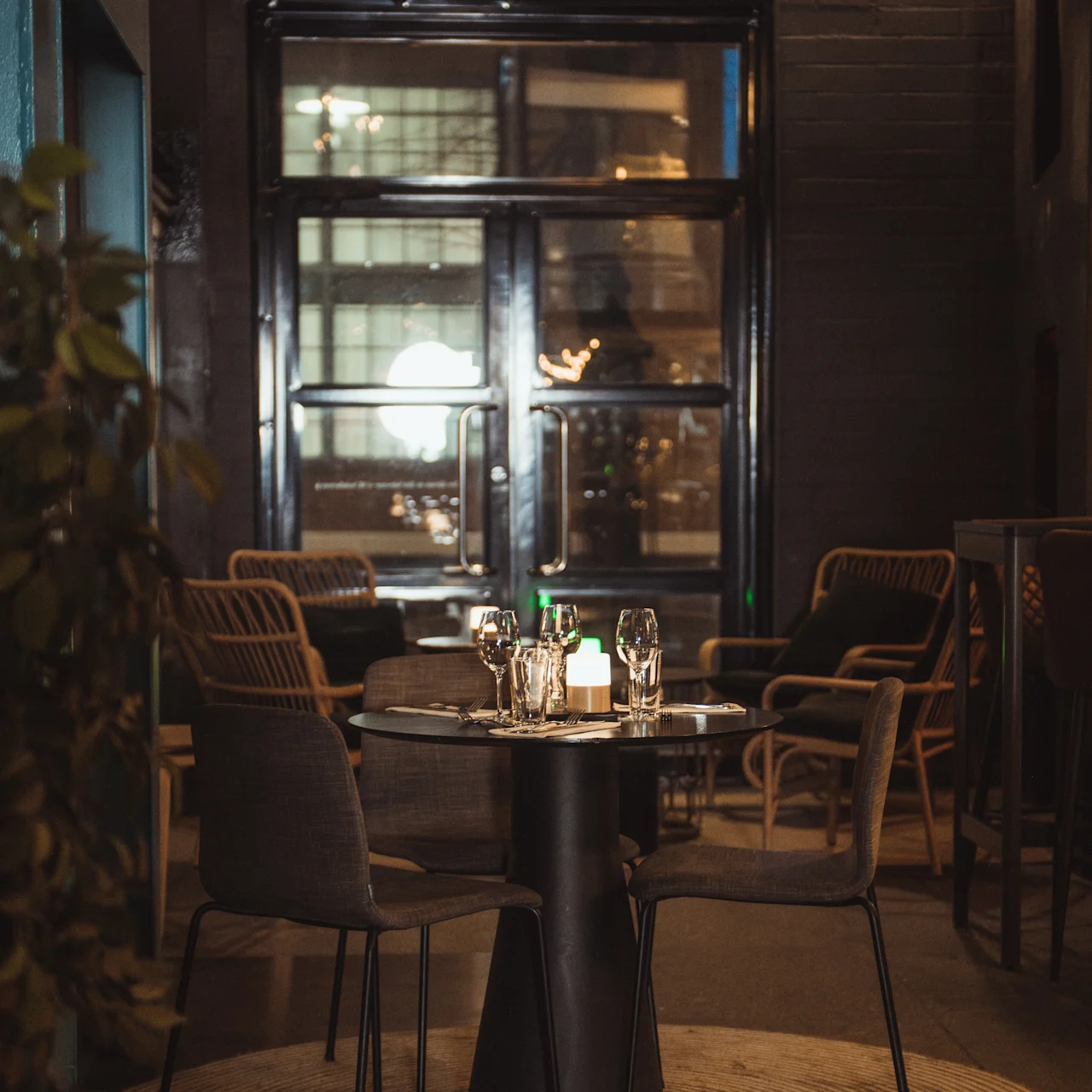 A round table set with glasses and cutlery stands under warm lighting in a cozy, modern café with wicker chairs and a large window in the background.