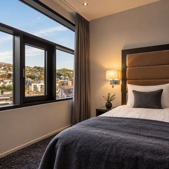 Koselig hotellrom med en myk seng, mørkeblått pledd, sengelampe og et panoramavindu med utsikt over byen.