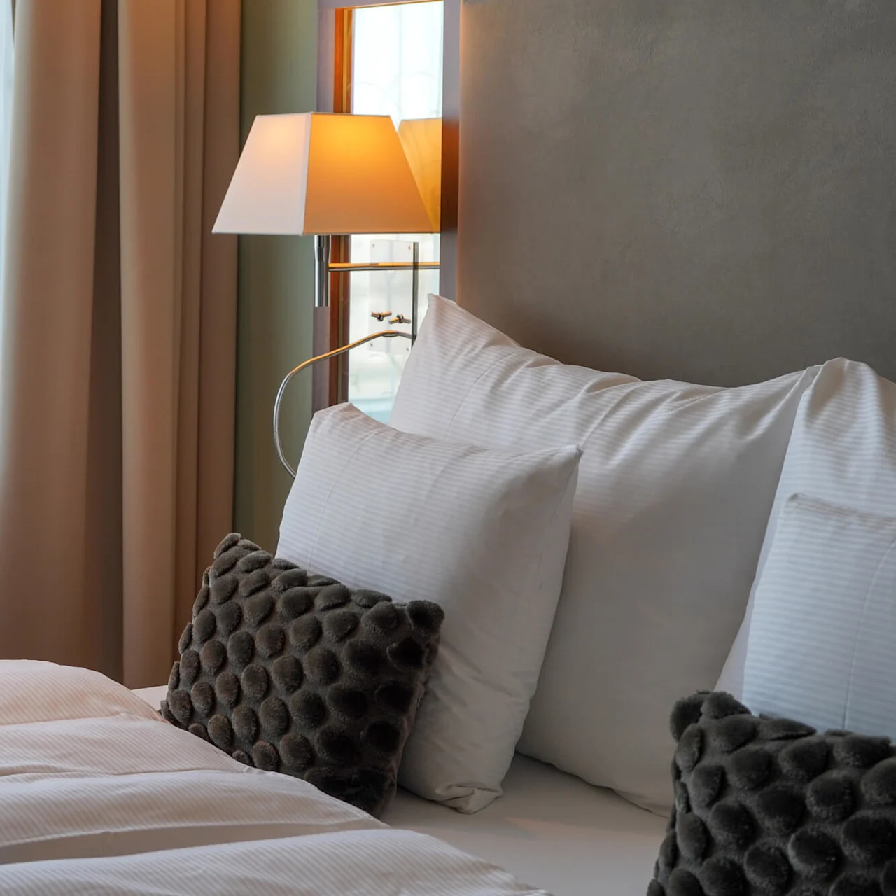 A neatly made bed features white pillows and textured dark cushions, beneath a warm-lit wall lamp. Nearby, sheer curtains cover a sunlit window.