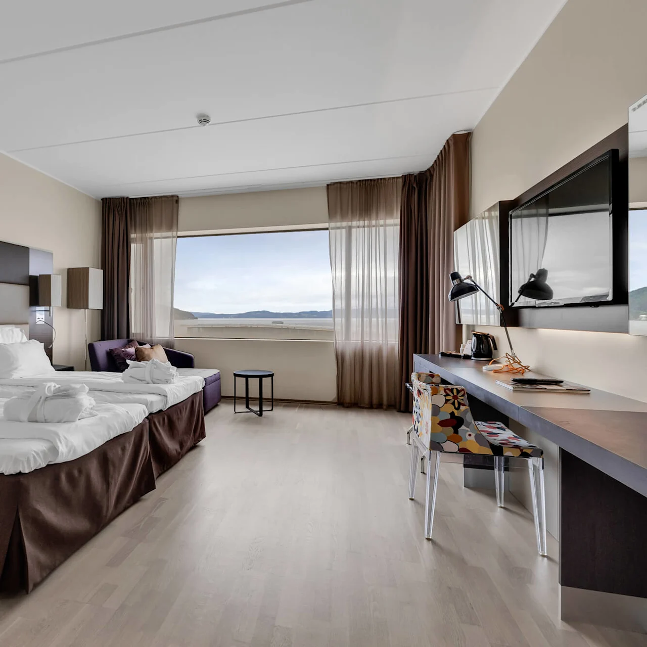 A king-size bed with white linens faces a wall-mounted TV in a modern hotel room with wooden floors. Large window with sheer curtains reveals a scenic view of hills.
