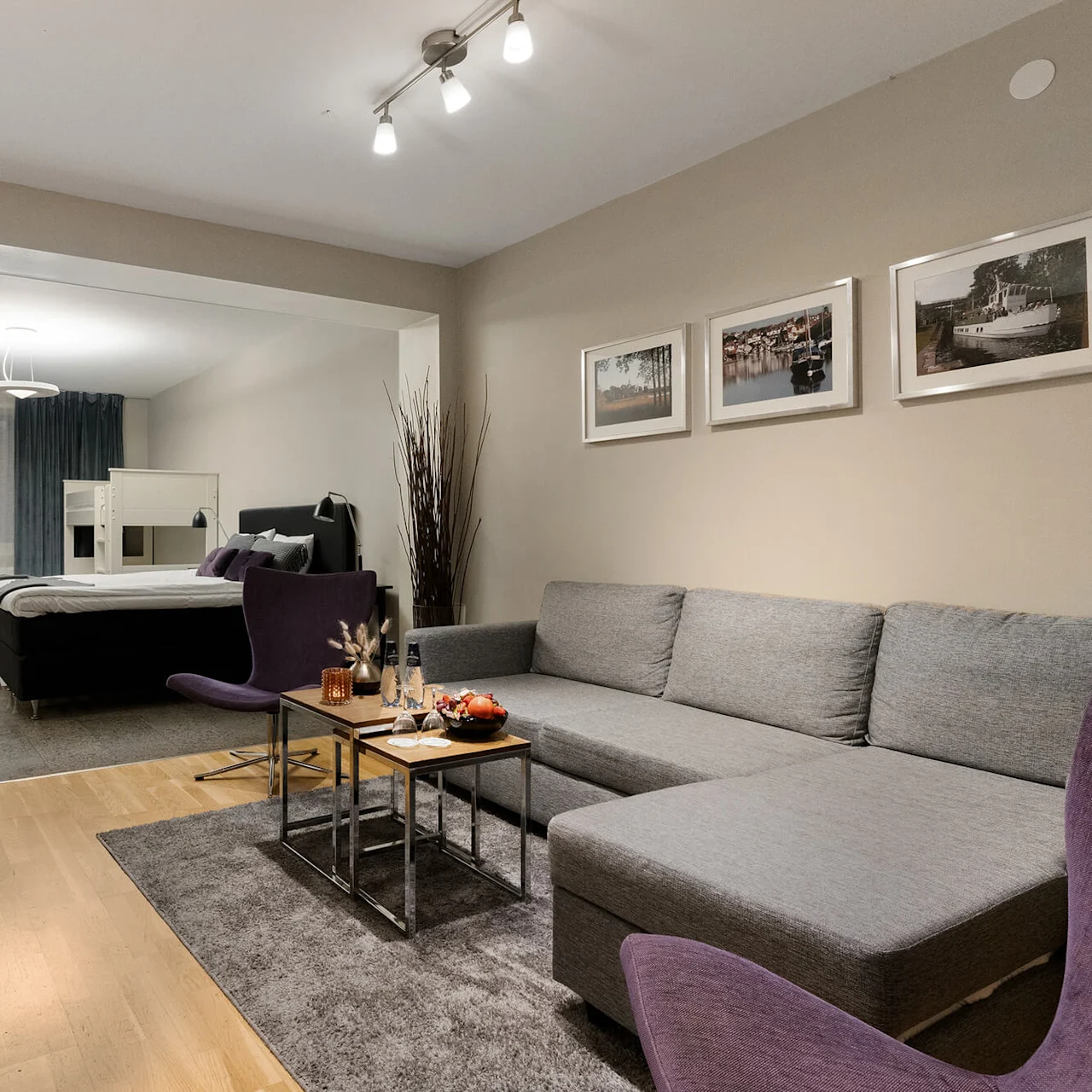 En grå hjørnesofa står i en stue vendt mod et lille bord med snacks, ved siden af et soveværelse med en seng. Indrammede fotografier pryder de beige vægge.
