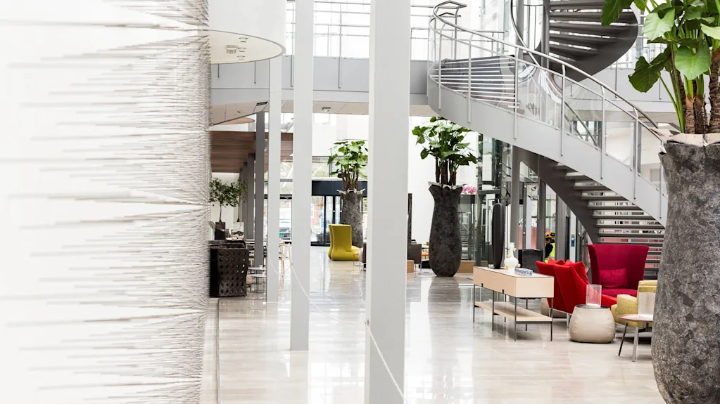 Large vases with plants decorate a modern atrium featuring a sleek, spiral staircase and colorful seating arrangements. Bright natural light filters through expansive windows, creating a spacious, contemporary ambiance.