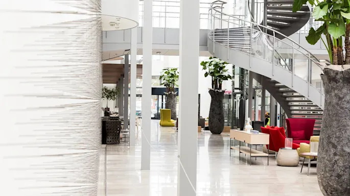 Large vases with plants decorate a modern atrium featuring a sleek, spiral staircase and colorful seating arrangements. Bright natural light filters through expansive windows, creating a spacious, contemporary ambiance.