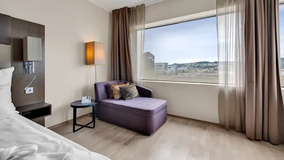 A purple chaise longue with pillows, positioned by a large window overlooking distant buildings, next to a side table with a card in a modern, light-filled room.