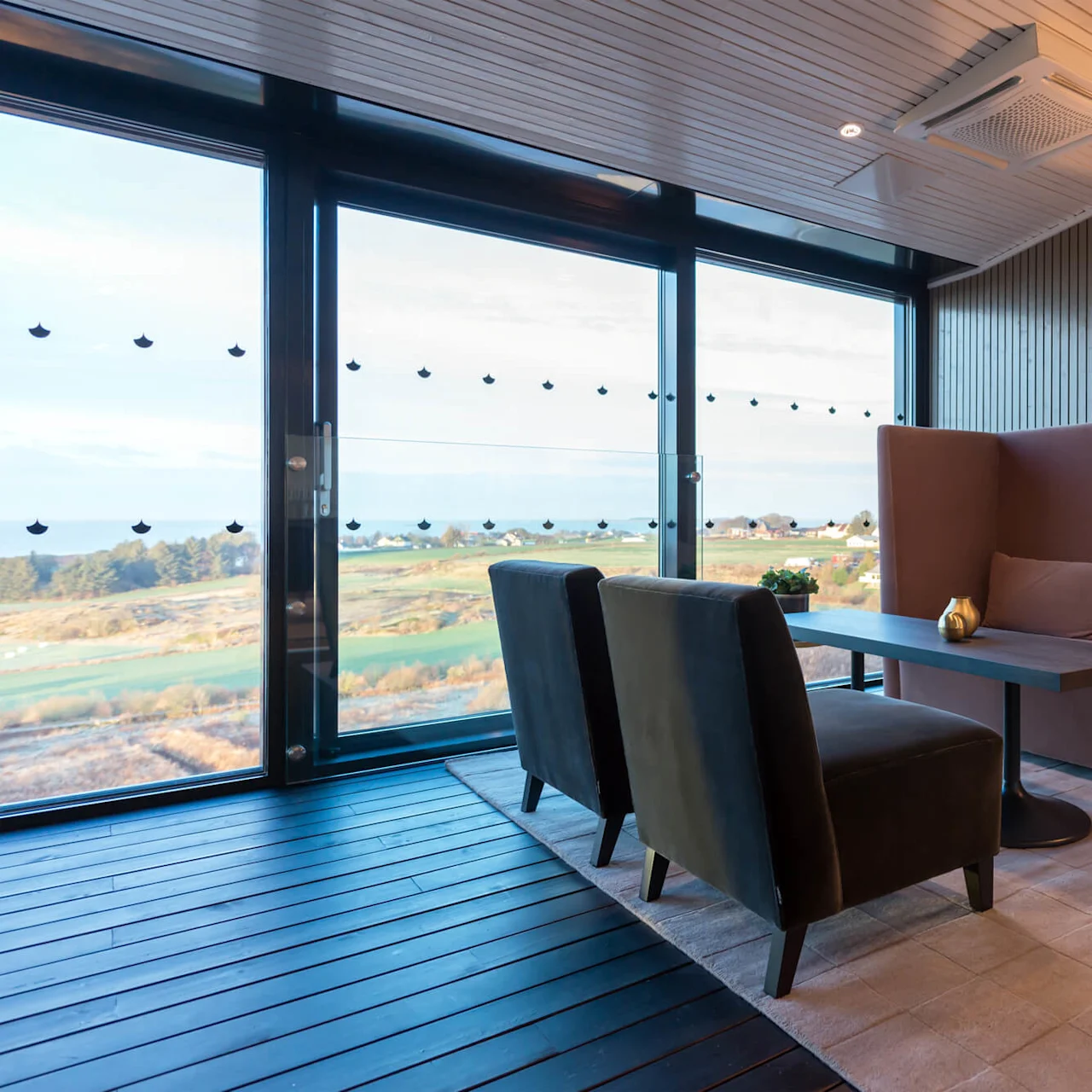 En koselig sittegruppe med en lang sofa, to lenestoler, et bord og en lampe. Store vinduer gir utsikt over et grønt landskap utenfor.
