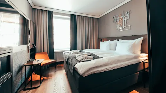 Seng med hvitt sengetøy står i et moderne rom, flankert av et lite bord og en oransje stol. Et vindu lyser opp det minimalistiske interiøret, med en veggskulptur over.