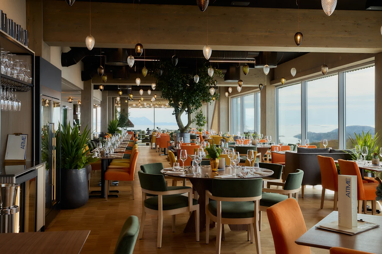 Restaurant med elegant dækkede borde, stole i grøn og orange, og store vinduer, der viser en naturskøn udsigt. Hængende lamper fra loftet forstærker den hyggelige atmosfære. Synlig tekst: "AIME".