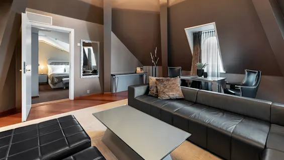 En moderne stue med en sort lædersofa og et glasbord, der forbinder til et soveværelse. Sollys strømmer ind gennem et vindue og fremhæver elegante møbler og neutrale toner.