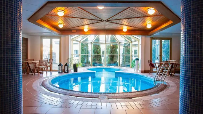 Et rundt innendørs basseng reflekterer taklys i et lyst rom med tremøbler, store vinduer og planter langs kantene.