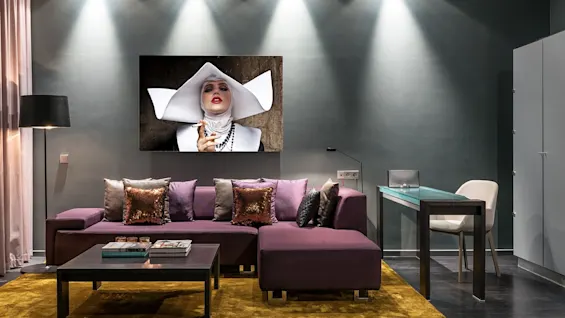 A modern living room features a purple sectional sofa adorned with pillows, facing a large, framed portrait of a fashionably dressed figure. Overhead lights illuminate the space, complemented by minimalist decor.