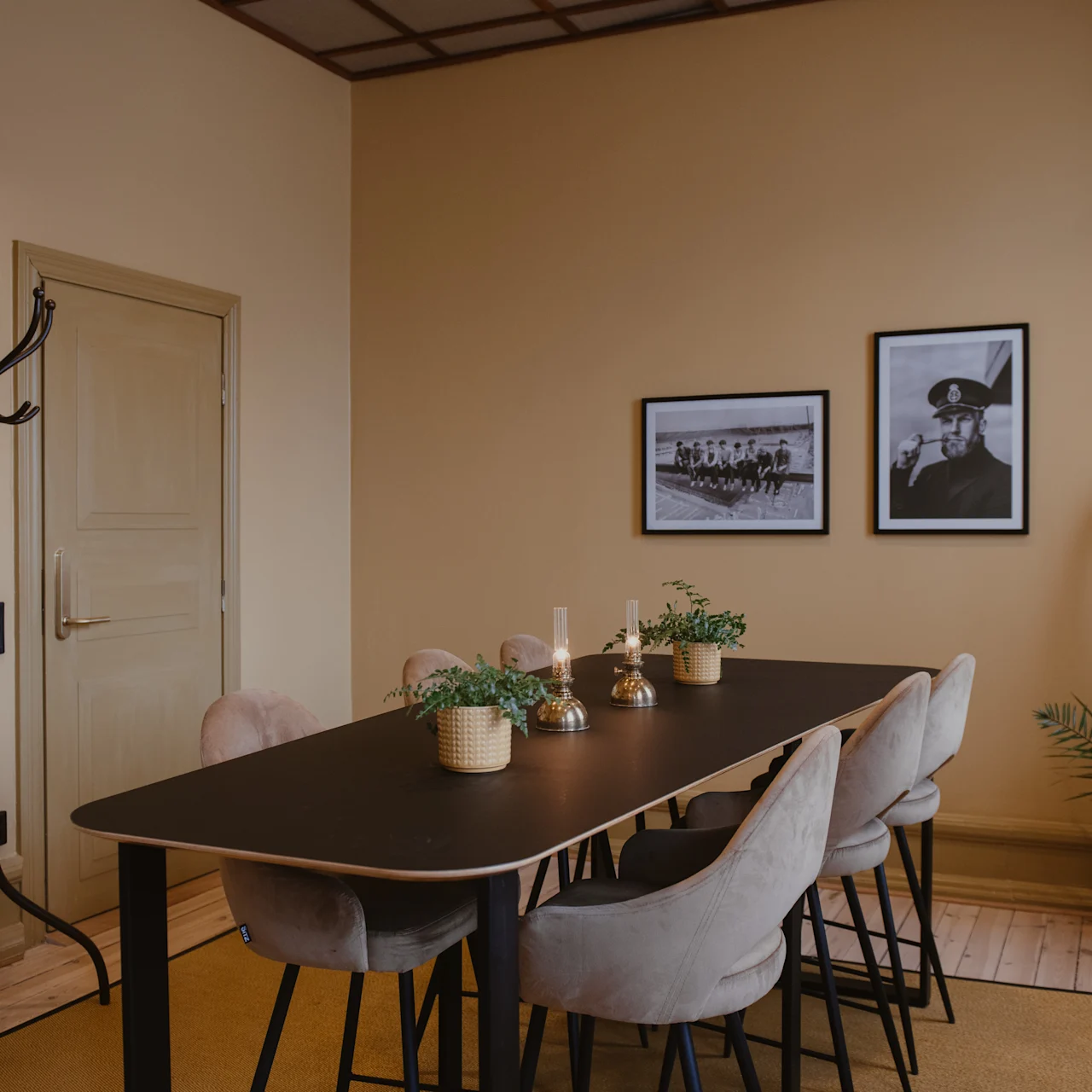 Ruokapöytä pehmeillä tuoleilla on koristeltu ruukkukasveilla ja kynttilöillä, sijoitettuna lämpimästi valaistuun huoneeseen, jossa on beige seinät, valokuvia kehyksissä ja naulakko lähellä.