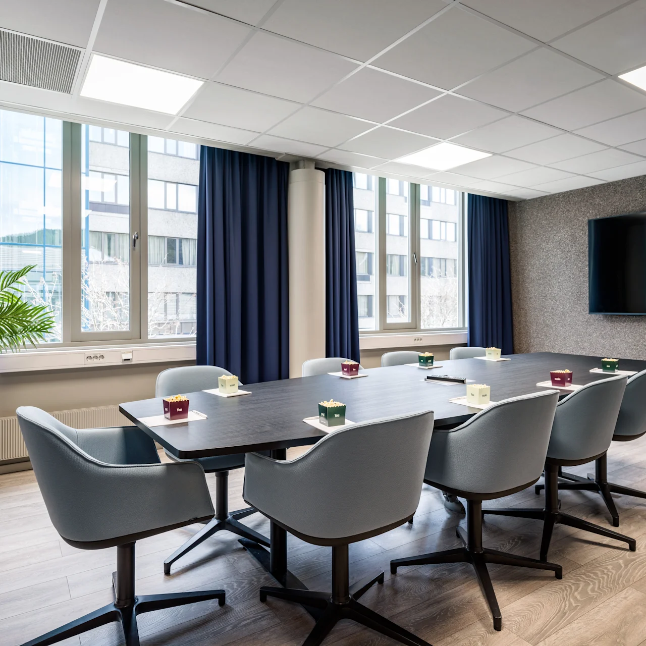 Eight gray chairs surround a rectangular conference table with coffee mugs. A plant sits by large windows with blue curtains, and a wall-mounted TV enhances the modern meeting room.