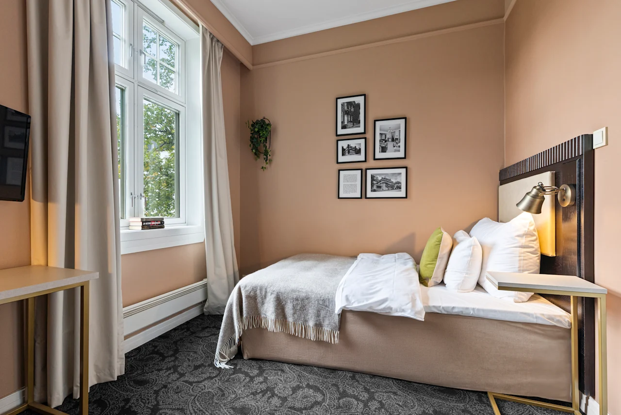 Et koselig hotellrom med en komfortabel seng, et stort vindu og innrammet kunst på veggen, som skaper en innbydende atmosfære.