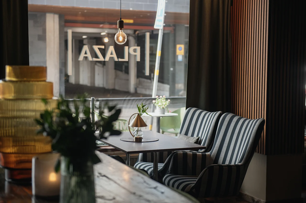 En mysig lounge med randiga fåtöljer vid ett fönster med texten "PLAZA." Ett bord har en liten lampa och växt, omgivna av dämpad belysning och dekorativa element som en glödlampa och vaser.