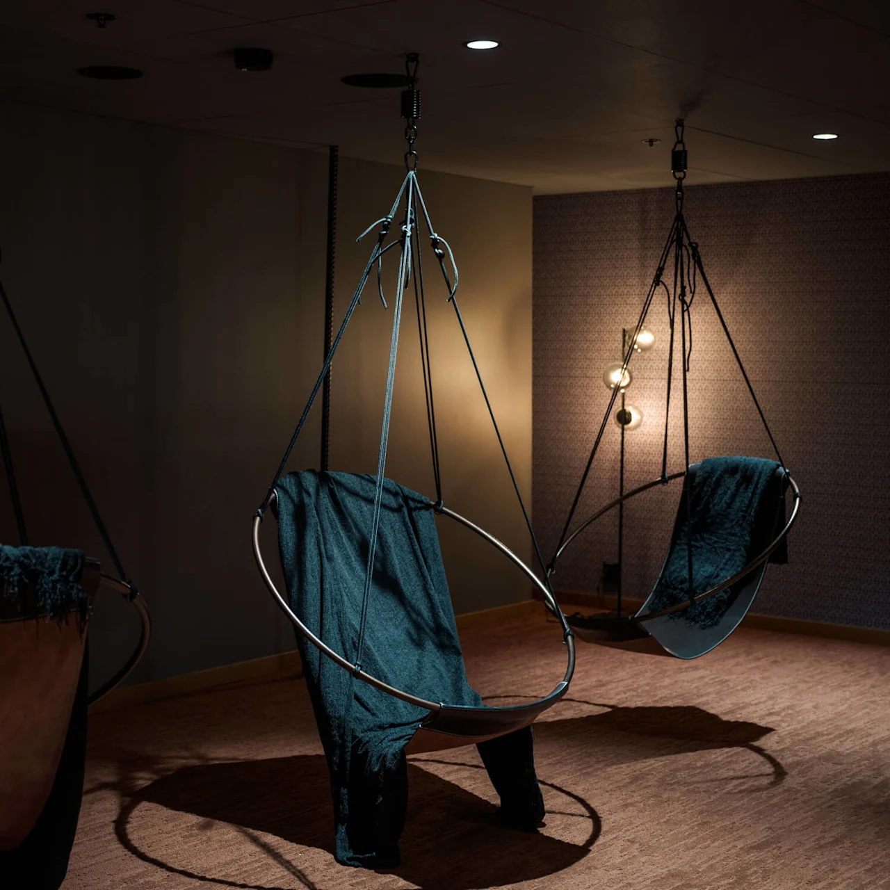 Hengestoler hengt fra taket, dekket med mørkt stoff, i et svakt opplyst rom med en teksturert vegg og en lampe med flere lyspærer i hjørnet.