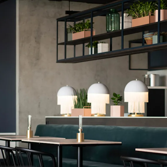 Bord med lampor i guldställ är placerade i ett modernt kafé. Ovanför finns hyllor med växter i rosa lådor mot en betongvägg, vilket tillför grönska till den stilrena miljön.