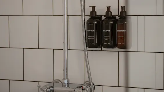 Mörka flaskor märkta "BODY WASH", "SHAMPOO" och "CONDITIONER" är monterade på en duschvägg ovanför en metallisk armatur, omgivna av vita kakelplattor.