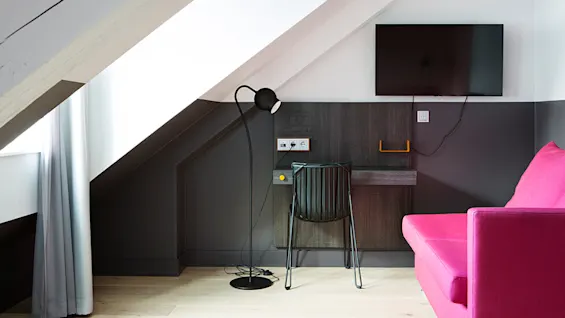 En svart stol og gulvlampe står ved et lite skrivebord under en veggmontert TV. Rommet har et skråtak, grå gardiner og en lys rosa sofa.