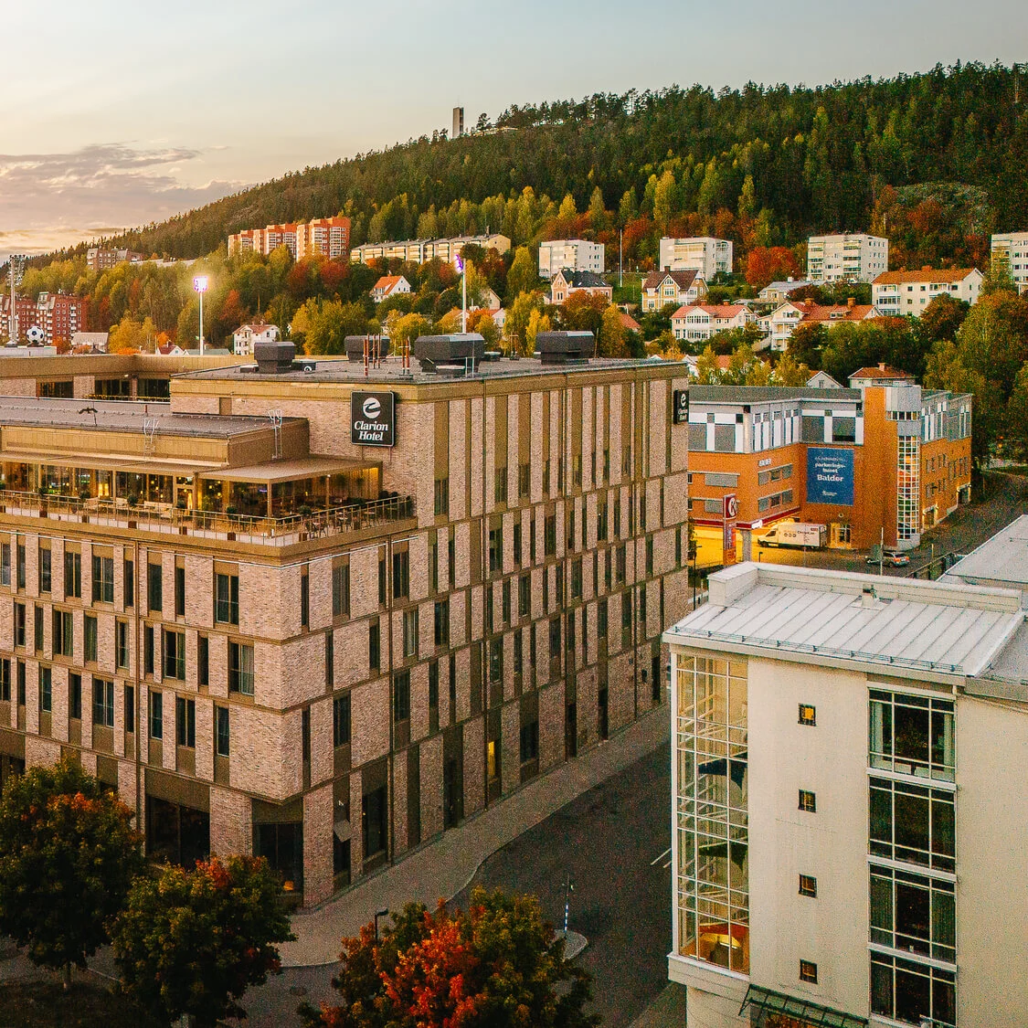En stor murstensbygning med "Clarion Hotel"-skilt er placeret midt i en by omringet af træer og andre bygninger under solnedgang. En skråning med huse er synlig i baggrunden.