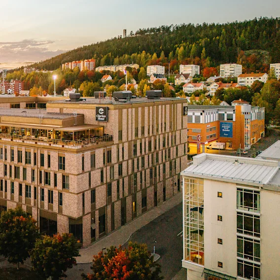A large brick building with "Clarion Hotel" signage is set in a cityscape surrounded by trees and other buildings during sunset. A hillside with houses is visible in the background.